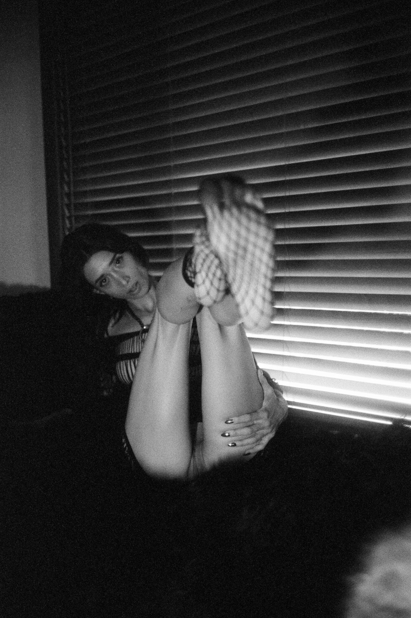 Black and white portrait, figure curled on a couch with light from venetian blinds casting striped shadows. Foot outstretched in fishnet stockings, hand holding the back of her thigh.