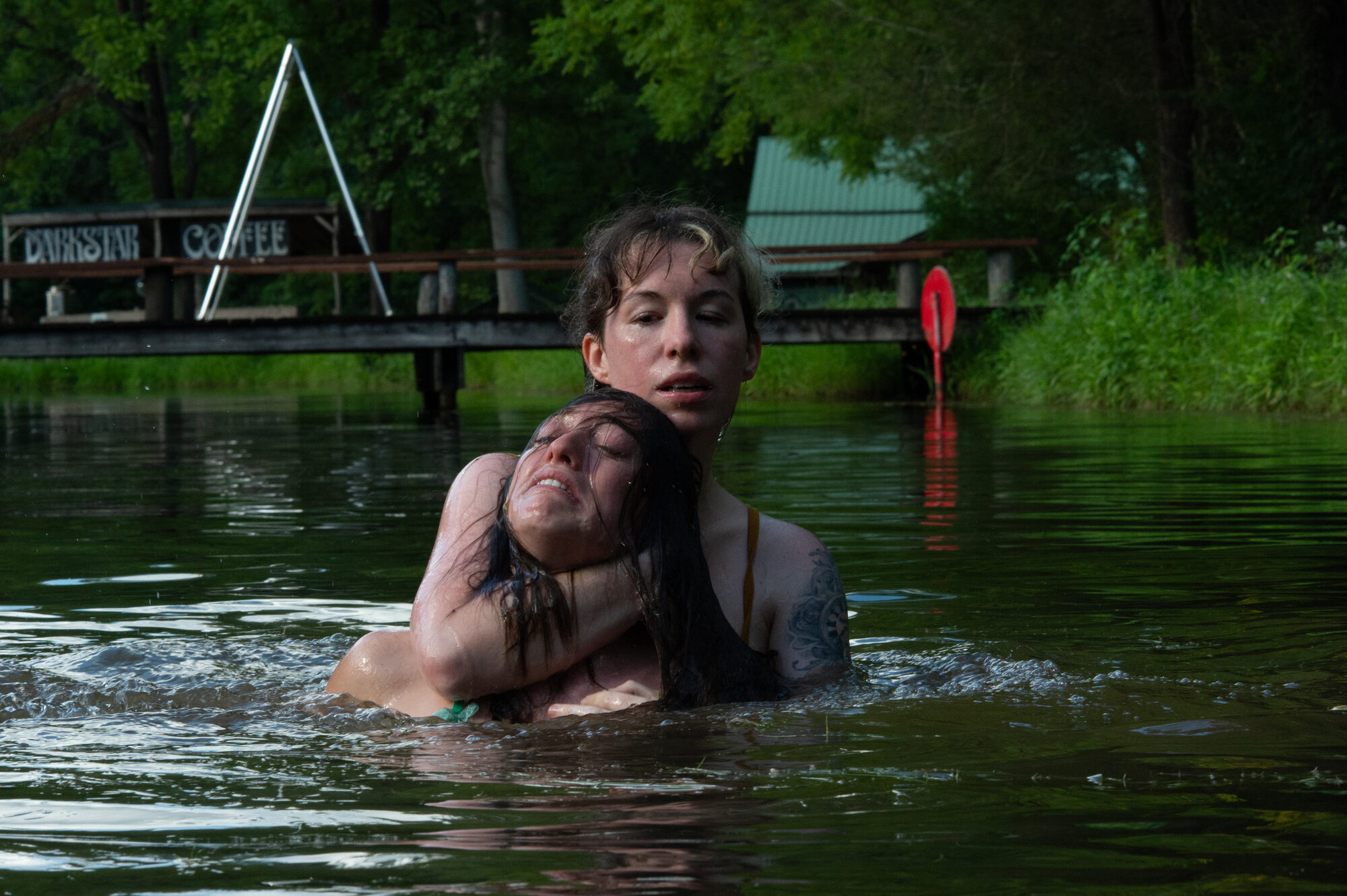 Two figures embracing in a green lake, one cradling the other, with a dock and trees behind. A consensual drowning scene with the bottom's legs tied in an agura.