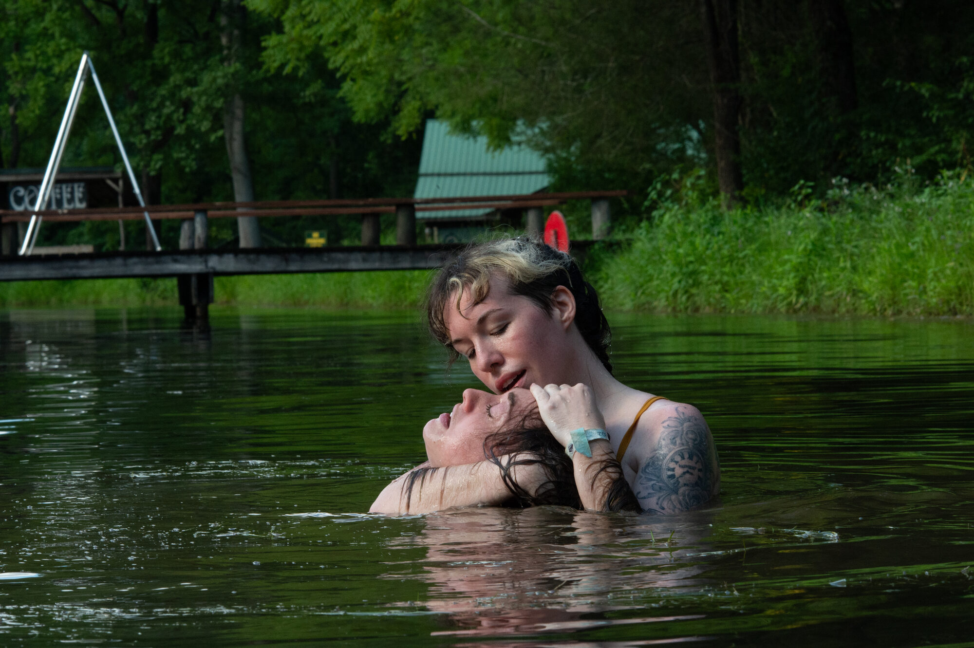 Two figures in a lake, one gently holding the other's face above water, eyes closed. A consensual drowning scene with the bottom's legs tied in an agura.