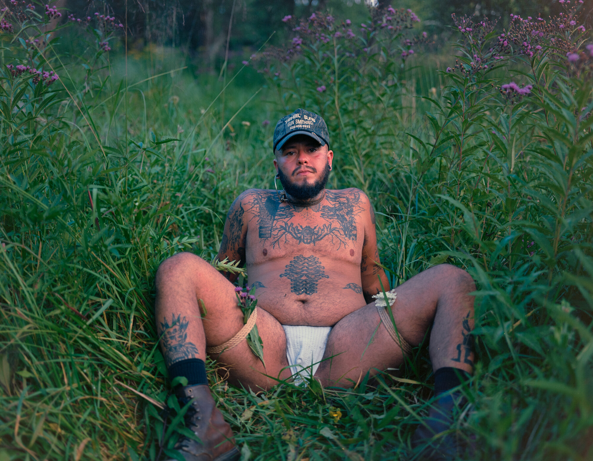Mo sits in a meadow of tall grass and purple ironweed, facing the camera in a camo cap, jockstrap, and boots, rope tied around both thighs. Tattooed chest and arms, evening light. Shot on 120 film.